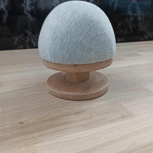 Op de afbeelding: Een grijze stoffen hoedvorm op een houten basis. De hoedvorm is rond en heeft een glad oppervlak. De basis is gemaakt van natuurlijk hout en heeft een ronde vorm.