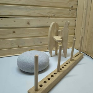May include: A light-colored wooden rack with three vertical pegs and a row of circular holes, a small light-colored wooden stool with a curved seat, and a round, light gray fabric-covered cushion arranged on a flat surface.
