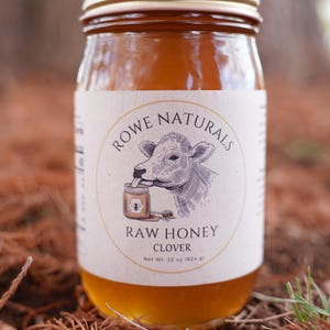 Puede incluir: Un tarro de cristal de miel de trébol cruda con tapa dorada. La etiqueta presenta una ilustración de vaca y el texto "Rowe Naturals". El tarro contiene 624 g de miel.