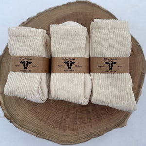 Puede incluir: Tres pares de calcetines de algod&oacute;n org&aacute;nico en diferentes tama&ntilde;os: peque&ntilde;o, mediano y grande. Los calcetines son de color crema natural y est&aacute;n doblados cuidadosamente sobre una superficie de madera. Cada par tiene una etiqueta marr&oacute;n con el texto "Run Naturals" y el tama&ntilde;o de los calcetines.
