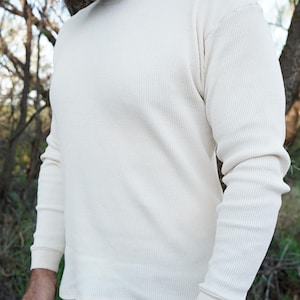 May include: A long-sleeve crew neck shirt in an off-white color, featuring a textured waffle-knit fabric pattern. The garment has a standard fit with ribbed cuffs at the wrists and a simple hemline at the waist, providing a clean and basic look.