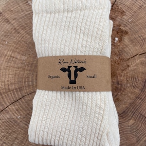 Puede incluir: Un par de calcetines blancos de algod&oacute;n org&aacute;nico doblados y enrollados. Los calcetines tienen una textura acanalada. Una etiqueta de papel marr&oacute;n con el texto "Rowe Naturals" y un logotipo de vaca est&aacute; unida a los calcetines. La etiqueta tambi&eacute;n dice "Organic", "Small" y "Made in USA".