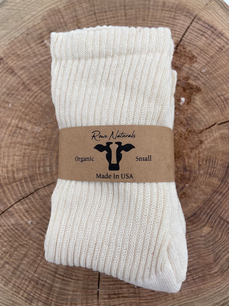Puede incluir: Un par de calcetines blancos de algod&oacute;n org&aacute;nico doblados y enrollados. Los calcetines tienen una textura acanalada. Una etiqueta de papel marr&oacute;n con el texto "Rowe Naturals" y un logotipo de vaca est&aacute; unida a los calcetines. La etiqueta tambi&eacute;n dice "Organic", "Small" y "Made in USA".