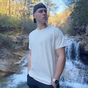 Op de afbeelding: Een jonge persoon draagt een wit T-shirt, een zwarte broek en een zwarte baseballpet en staat voor een waterval. De persoon kijkt naar rechts van het frame.