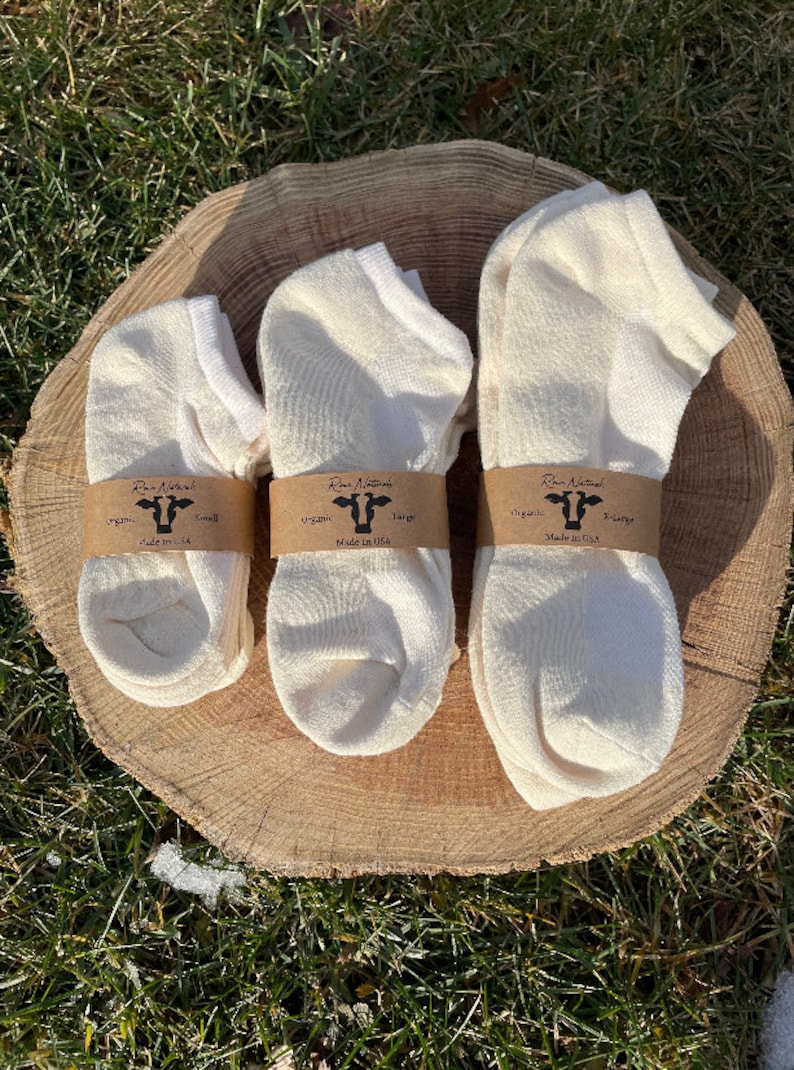 Puede incluir: Tres pares de calcetines org&aacute;nicos de color blanquecino en diferentes tallas, peque&ntilde;a, grande y X-grande, se muestran sobre una rodaja de madera. Cada par est&aacute; envuelto con una banda de papel marr&oacute;n con un gr&aacute;fico de vaca y el texto "Raw Naturals, organic, Made in USA."