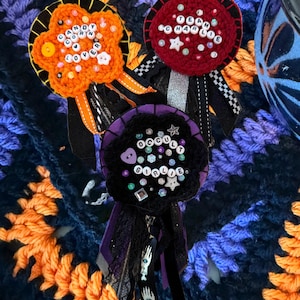 May include: Three handmade circular fabric rosettes featuring orange, red, and black crochet centers. The rosettes are decorated with beads, buttons, and letter beads spelling "CANDY CORN LOVER," "TEAM CHARLIE," and "OCCULT GIRLIE," with dangling ribbons.