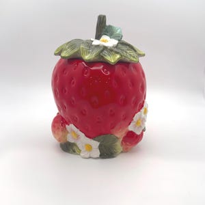 May include: A ceramic jar shaped like a large red strawberry with a textured surface. The lid features a green leafy stem and a small white flower. Additional white flowers with yellow centers and green leaves decorate the base of the jar.