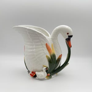 Può includere: Brocca a forma di cigno in ceramica bianca, con un lungo collo curvo e un manico. La brocca presenta un design dettagliato di piume, con accenti arancioni e verdi. Una piccola statuina di cigno è alla base, con dettagli floreali rossi e verdi.