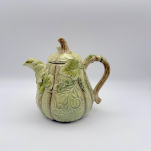 Puede incluir: Tetera de cerámica con forma de calabaza, de color verde claro con detalles marrones. La tetera tiene tapa, asa y boquilla, con diseños detallados de hojas y vides. Un artículo decorativo.
