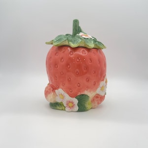 May include: A ceramic jar shaped like a large red strawberry with a green stem lid. The base features sculpted white strawberry blossoms with yellow centers and green leaves. The surface has a glossy finish with textured seed indentations.