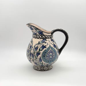 May include: A ceramic pitcher featuring a cream-colored base decorated with intricate blue and green floral patterns, including tulip motifs and a central teardrop design. The handle is solid black, and the base and rim are accented with black and white scrollwork.