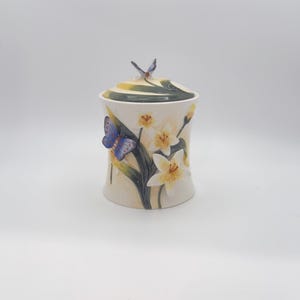 May include: A cylindrical ceramic jar with a lid featuring a small butterfly finial. The jar is decorated with raised, three-dimensional white lily flowers, green foliage, and a prominent blue and purple butterfly on the side of the vessel.