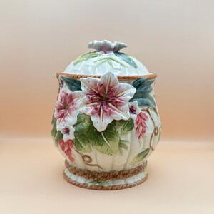 Può includere: Un barattolo in ceramica con coperchio caratterizzato da un design floreale in rilievo tridimensionale. Il barattolo ha una base color crema con scanalature verticali, decorato con fiori rosa e bianchi, foglie verdi e un bordo a corda.