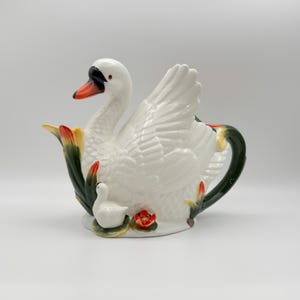 Puede incluir: Tetera de cerámica blanca con forma de cisne, con pico negro y detalles en naranja. La tetera presenta un cisne con las alas extendidas, un cisne pequeño y detalles florales. El asa es de color verde oscuro.