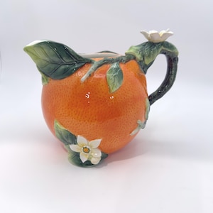 May include: A ceramic pitcher shaped like a textured orange, featuring a dark green handle, green leaves, and white blossoms. The surface has a realistic dimpled orange peel texture, with a glossy finish and a spout integrated into the fruit design.