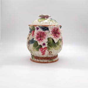 May include: A ceramic canister with a lid featuring a cream-colored base with a vertical ribbed texture. The exterior is decorated with raised, three-dimensional pink and white flowers, green leaves, and a brown rope-like trim at the base.