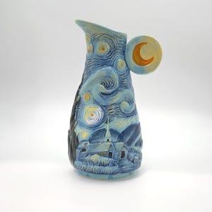 May include: A ceramic pitcher featuring a hand-painted blue and yellow night sky scene with swirling clouds, stars, and a crescent moon on the handle. The base depicts a village landscape with a church steeple in shades of blue and dark tones.