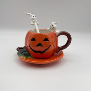 May include: A ceramic Halloween-themed teacup and saucer set. The orange teacup is shaped like a jack-o'-lantern with a smiling face. A white ghost spoon rests inside the cup, and a ghost figure is perched on the rim. The saucer is orange with green leaf accents.