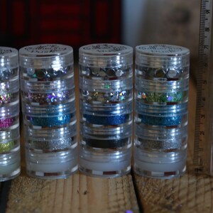 May include: Four clear plastic containers with multiple layers of glitter and sequins. Each container has a label that reads "The Glitter Push".