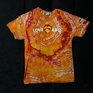 Pode incluir: Camiseta tie-dye laranja e vermelha com o texto "LOVE ABQ" e um gráfico de coração. O texto "#loveabq" também está incluído.