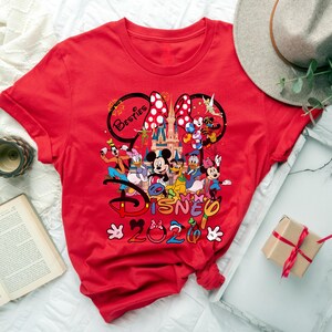 May include: A red t-shirt featuring a colorful graphic of Disney characters, including Mickey and Minnie Mouse, Donald Duck, and Goofy. The design includes the text "Besties" and "Disney 2020". The shirt has a crew neck and short sleeves.