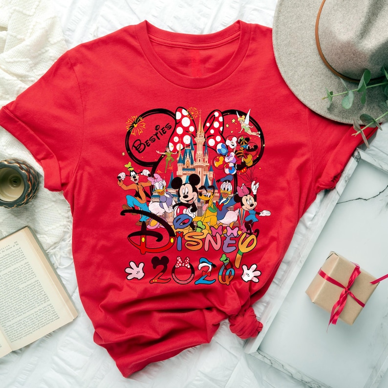 May include: A red t-shirt featuring a colorful graphic of Disney characters, including Mickey and Minnie Mouse, Donald Duck, and Goofy. The design includes the text "Besties" and "Disney 2020". The shirt has a crew neck and short sleeves.