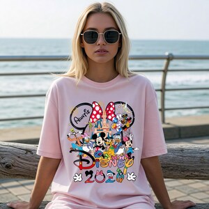 May include: A light pink t-shirt featuring a colorful Disney graphic. The graphic includes Mickey and Minnie Mouse, Donald Duck, and other characters, with the text "Disney 2020" and "Auntie".