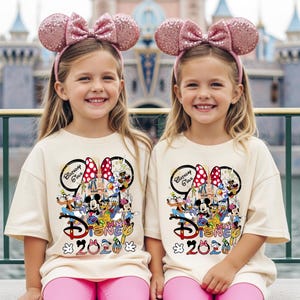 May include: Two young girls wearing matching pink sequined Minnie Mouse ear headbands and cream-colored t-shirts with a Disney 2020 graphic featuring Mickey Mouse and friends. They are also wearing pink pants.