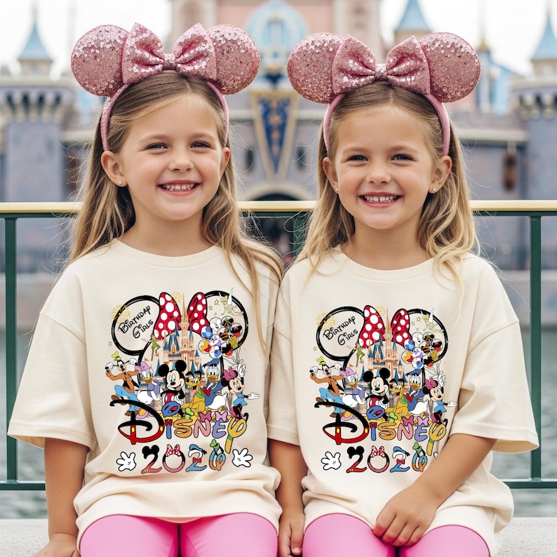 May include: Two young girls wearing matching pink sequined Minnie Mouse ear headbands and cream-colored t-shirts with a Disney 2020 graphic featuring Mickey Mouse and friends. They are also wearing pink pants.
