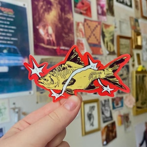 May include: A die-cut sticker featuring a yellow fish illustration with black line details, surrounded by a bright red border and accented with white four-pointed star shapes that appear to be piercing through the body of the fish.