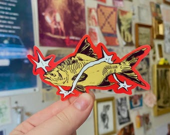 May include: A die-cut sticker featuring a yellow fish illustration with black line details, surrounded by a bright red border and accented with white four-pointed star shapes that appear to be piercing through the body of the fish.