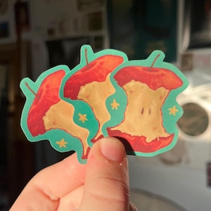 May include: Three red apple core stickers on a teal background. Each apple core has a yellow center and a red rim. There are small yellow stars scattered on the background.