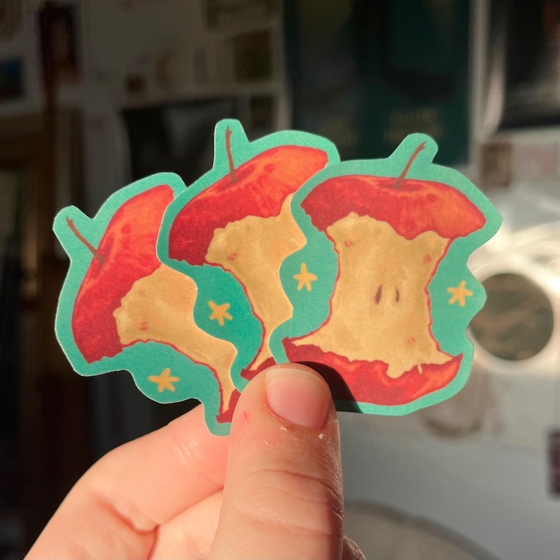 May include: Three red apple core stickers on a teal background. Each apple core has a yellow center and a red rim. There are small yellow stars scattered on the background.