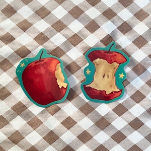 May include: Two red apple stickers, one whole and one with a bite taken out. The stickers are on a brown and white checkered background.