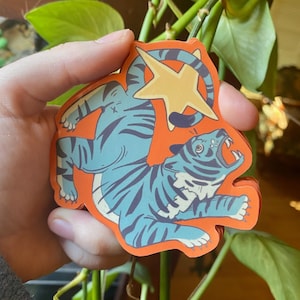 Könnte beinhalten: Ein Aufkleber mit einem stilisierten blauen Tiger mit schwarzen Streifen auf orangefarbenem Hintergrund. Der Tiger hat ein offenes Maul und einen gelben Stern darüber. Der Aufkleber wird in einer Hand gehalten.