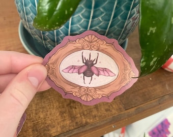 May include: A pink and brown beetle with wings is depicted in a decorative oval frame with gold accents. The frame has a pink background and is surrounded by a floral design.