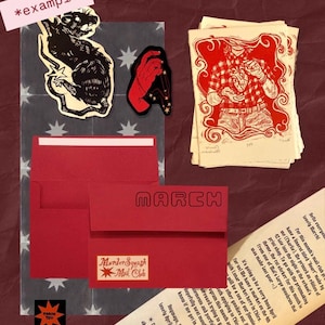 Puede incluir: Una colecci&oacute;n de art&iacute;culos de papeler&iacute;a que incluye un sobre rojo con el texto MARCH y MurderSquash Mail Club, una pegatina de rata negra, una pegatina de mano roja, una pila de impresiones de corazones rojos y blancos y una carta.