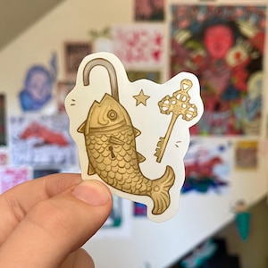 Può includere: Un adesivo con un design stravagante di un lucchetto a forma di pesce con un grillo aperto, una stella e una chiave. Il lucchetto a pesce è color oro con dettagli di squame e la chiave è anch'essa dorata. L'adesivo è su sfondo bianco.