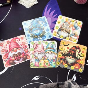 May include: A selection of square gift cards featuring gnome illustrations. The cards showcase various themes, including birthdays and holidays. Each card has a different design, with colorful illustrations and text. Price tags are visible.
