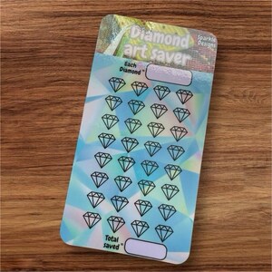 May include: A blue and white diamond art saver tracker with a holographic finish. The tracker has a space to write the number of diamonds used per project and the total number of diamonds saved. The tracker has 49 diamond icons.