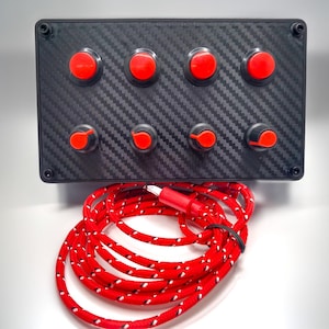 Könnte beinhalten: Eine schwarze Steuerung mit Carbonfaserstruktur hat acht rote Knöpfe und Drehregler. Ein rot-weiß geflochtenes Kabel ist mit dem Panel verbunden. Das Panel ist für elektronische Geräte konzipiert.