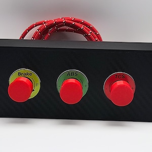 May include: A black control box with three red push buttons labeled "Brake", "ABS", and "TCS". The buttons are set in green and red circles. A red and white braided cable extends from the top of the box.