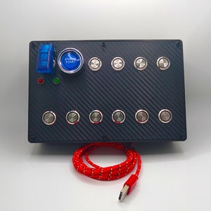 Può includere: Un pannello nero con una texture in fibra di carbonio presenta una varietà di pulsanti, tra cui un pulsante rotondo rosso con la scritta "Avvio motore" e un interruttore a levetta blu. Un cavo USB rosso è collegato al pannello.