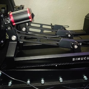 May include: Black SimuCUBE motion simulator with two black actuators and a red and black SimCore unit. The actuators are attached to a black metal frame with a black base. The SimCore unit is attached to the frame with a black metal bracket. The base has the word "SimuCUBE" printed on it.