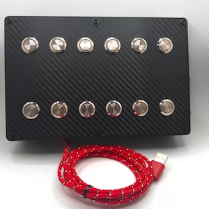 Peut inclure: Une boîte noire avec 10 boutons argentés disposés en deux rangées. La boîte a un motif en fibre de carbone et est connectée à un câble USB rouge.