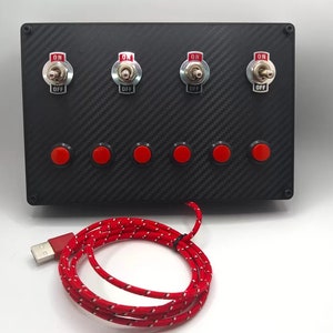 Pode incluir: Um painel preto com quatro interruptores de alternância etiquetados como "ON" e "OFF" e seis botões de pressão vermelhos. Um cabo USB vermelho está conectado ao painel.