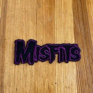 Puede incluir: Parche negro con la palabra "Misfits" en letras moradas estilizadas. El parche tiene un fondo negro con costuras moradas alrededor de las letras. El parche está sobre una superficie de madera.