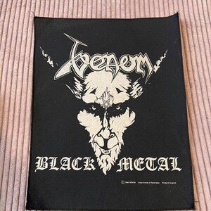 Può includere: Toppa quadrata nera con il nome della band "Venom" in un font stilizzato sopra un'illustrazione bianca di un volto cornuto. Le parole "BLACK METAL" sono sotto l'illustrazione in un font gotico.