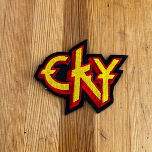 May include: Embroidered patch featuring the letters "CKY" in yellow, outlined in red, and set against a black background. The patch is in a stylized, angular font. The patch is on a wooden surface.