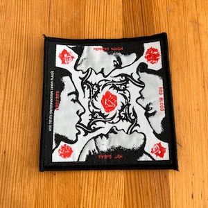 May include: Square black patch with a white and black design featuring four profiles and a central rose. The patch has red roses in each corner and text that reads "PEPPER'S MACK", "RED BLOOD", "HOT SUGAR", and "KISS-SEX".